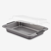 A rectangular non-stick baking pan with handles, featuring a transparent plastic lid slightly lifted above it. The lid is designed to securely cover the pan, providing convenience for storage or transport.