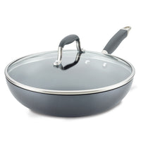 A gray non-stick frying pan with a clear glass lid and a black handle is shown. The lid has a metallic rim and handle. The pan is slightly angled, showcasing its deep design and shiny finish.