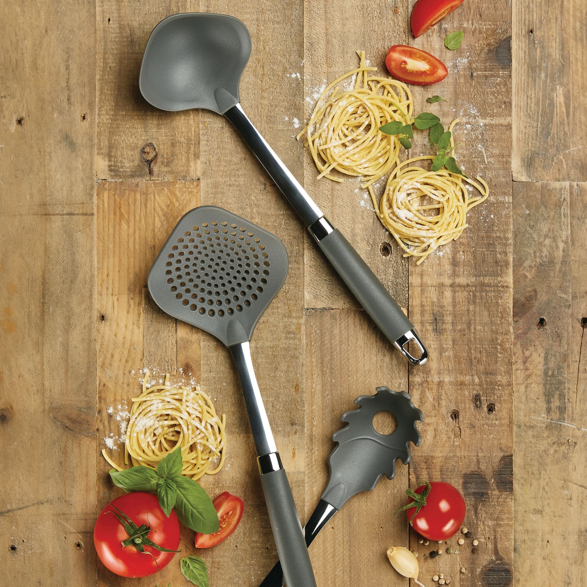 Three kitchen utensils on a wooden table surrounded by nests of uncooked spaghetti, cherry tomatoes, basil leaves, and scattered flour. The utensils are a ladle, a skimmer, and a pasta fork, all with gray heads and handles.