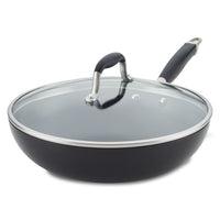 A large, black non-stick frying pan with a glass lid and black handles sits on a white surface. The metal rim around the glass lid contrasts with the smooth black finish of the pan.