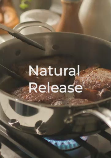 A 20-second video titled "Natural Release" featuring a chef in a pink shirt and apron. The video shows a close-up of thick, bone-in pork chops searing in an Anolon EverLast Clad Stainless Steel frying pan. As the meat browns, it demonstrates the "natural release" property of the tri-ply surface, with the pork chops lifting away effortlessly without sticking once a proper crust has formed.