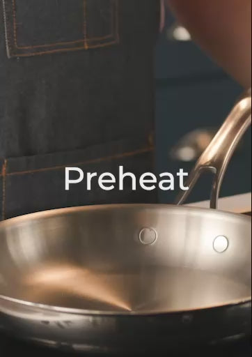 A 20-second instructional video titled "Preheat" featuring a chef in a dark apron. The video shows a close-up of an Anolon EverLast Clad Stainless Steel frying pan on a gas stove. Two water droplets are flicked into the pan; they bead up and dance across the surface (the Leidenfrost effect), indicating the pan is perfectly preheated for a natural nonstick release.