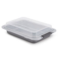 A rectangular gray baking pan with a detachable transparent plastic lid. The lid is securely fastened, and the brand name Anolon is featured on the handle. The pan is designed for baking and transporting food.