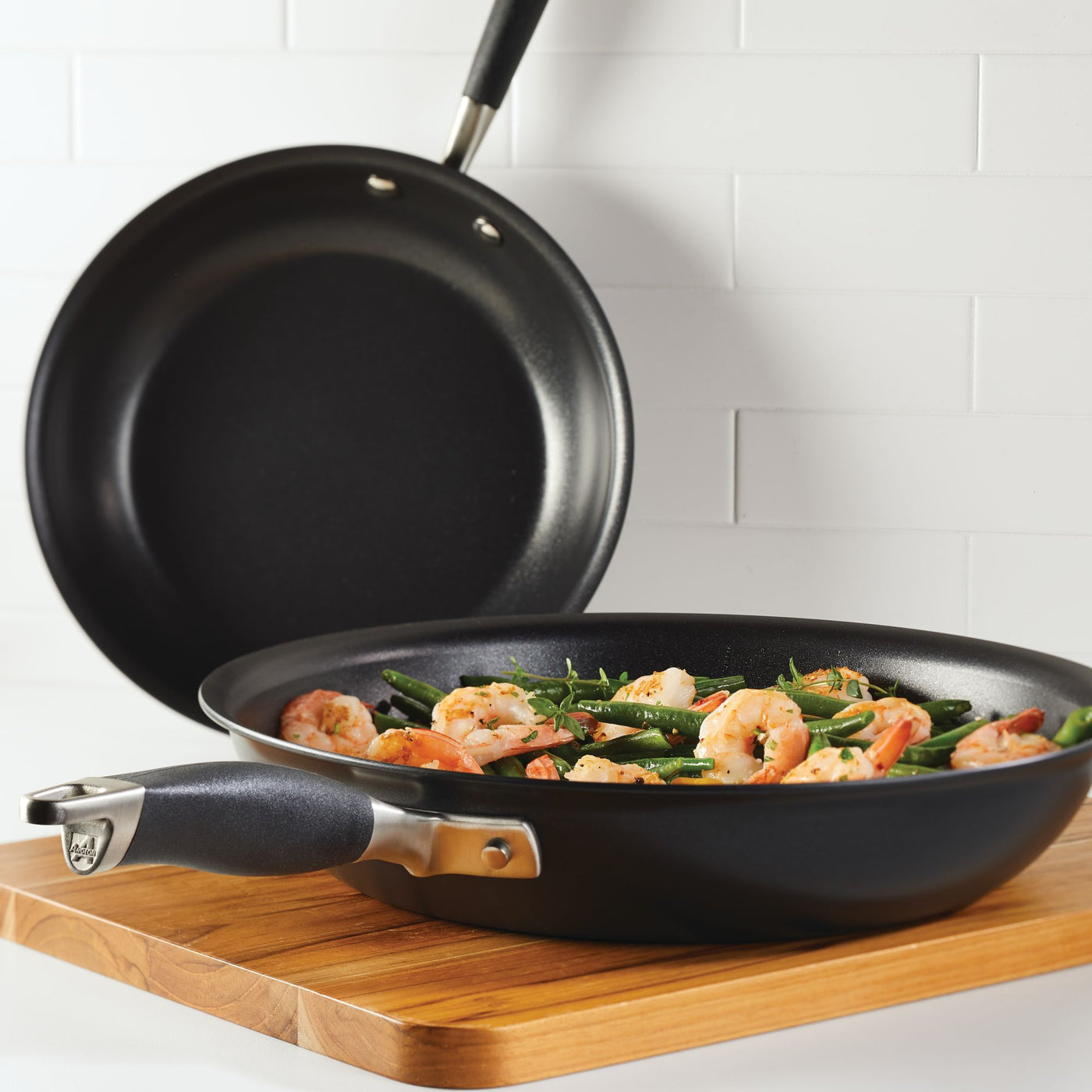 A frying pan filled with shrimp and green beans is placed on a wooden board. Another empty pan is leaned against white tiled wall in the background.