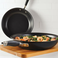 A frying pan filled with shrimp and green beans is placed on a wooden board. Another empty pan is leaned against white tiled wall in the background.