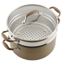 A metal steamer pot with a glass lid, featuring brown handles, is shown. The pot has a perforated insert for steaming and is placed on a solid base.