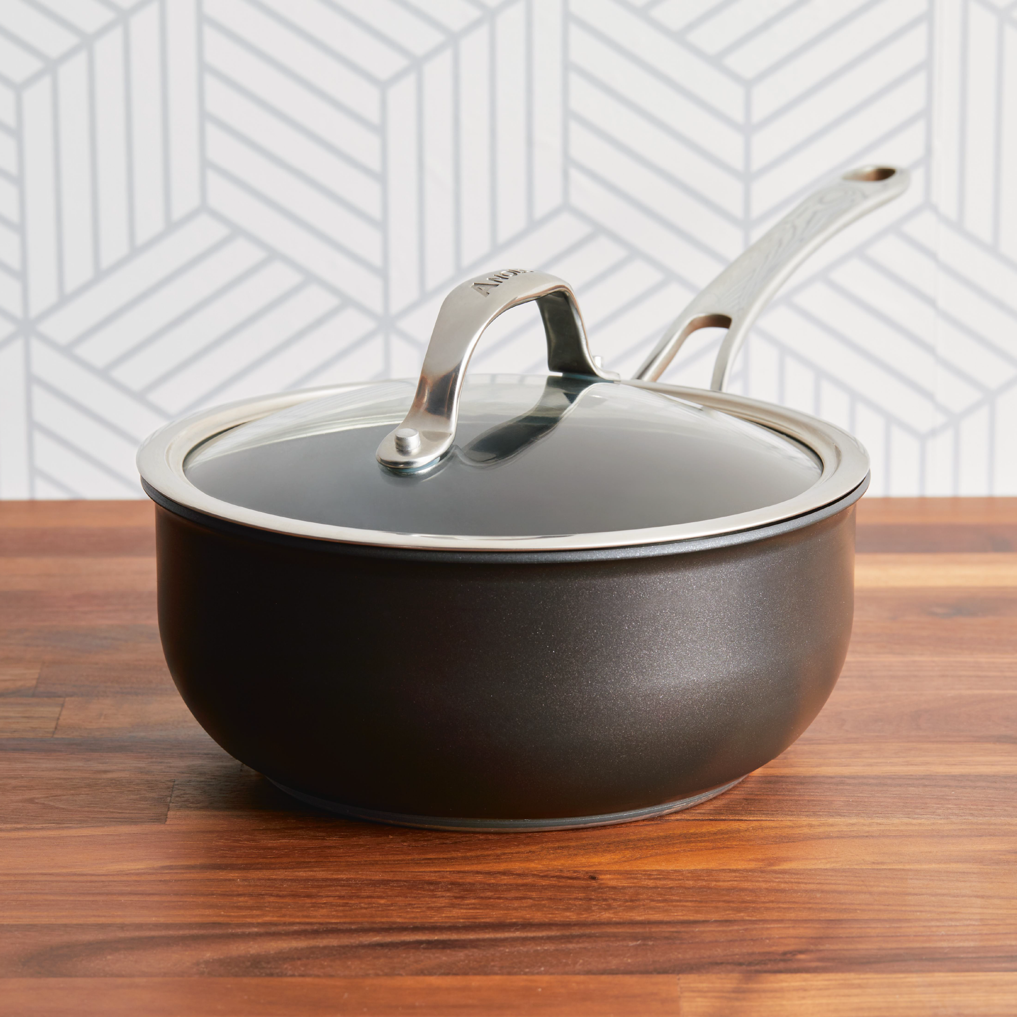 The Anolon X 2.5-Quart Hybrid Nonstick Saucier Pan, featuring a glass lid and metal handle, rests on a wooden surface with a geometric gray patterned wall in the background.