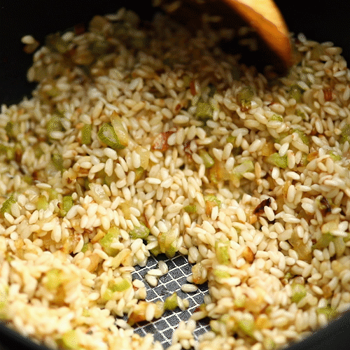 A close-up of a 2.5-Quart Hybrid Nonstick Saucier Pan by Anolon X, showcasing sizzling golden-brown rice mixed with chopped green bell peppers and onions. A wooden spoon stirs the mixture, revealing a grid-like pattern bottom without concern.
