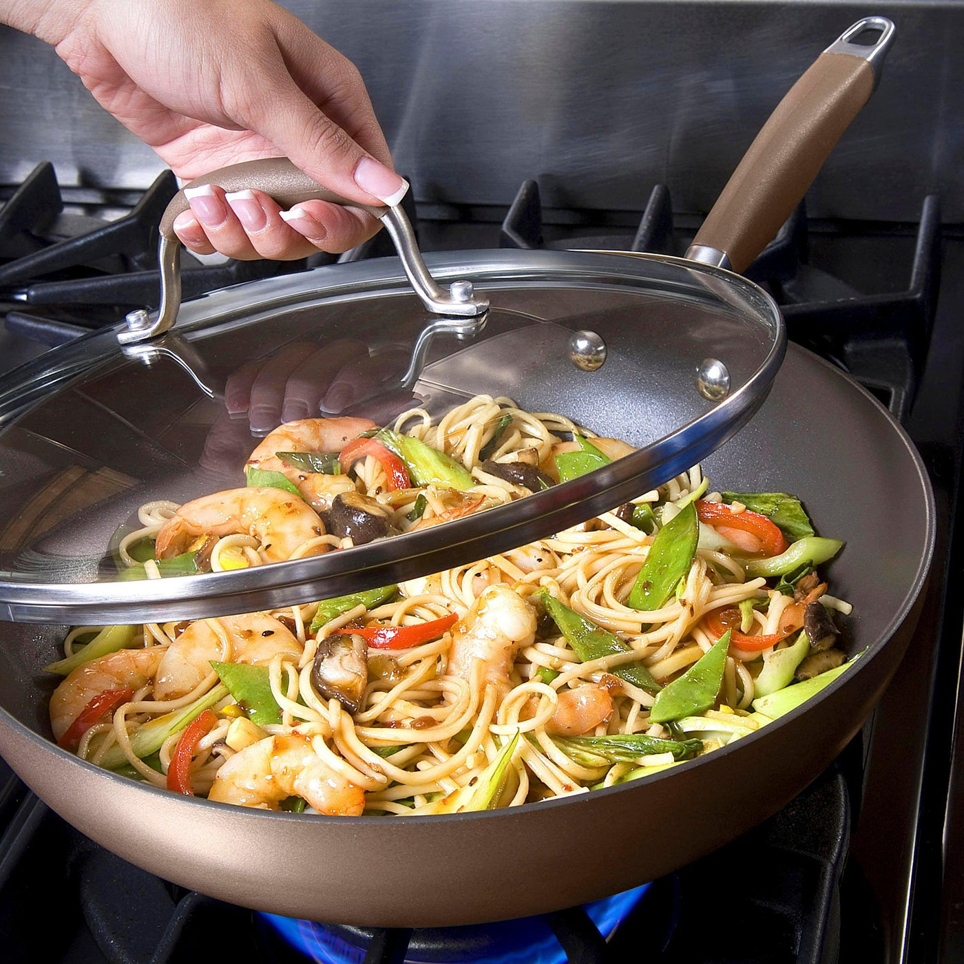 A hand lifts the glass lid of the Advanced 12-Inch Covered Ultimate Pan on a stove, revealing shrimp stir-fry with noodles, snap peas, mushrooms, and red bell peppers cooking inside.