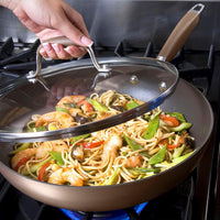 A hand lifts the glass lid of the Advanced 12-Inch Covered Ultimate Pan on a stove, revealing shrimp stir-fry with noodles, snap peas, mushrooms, and red bell peppers cooking inside.