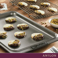 Chocolate-dipped cookies with chopped pistachios rest on the Advanced 11 x 17-Inch Baking Sheet and Cooling Rack Set, with a bowl of pistachios nearby. The Anolon logo appears at the bottom of this elegant scene.