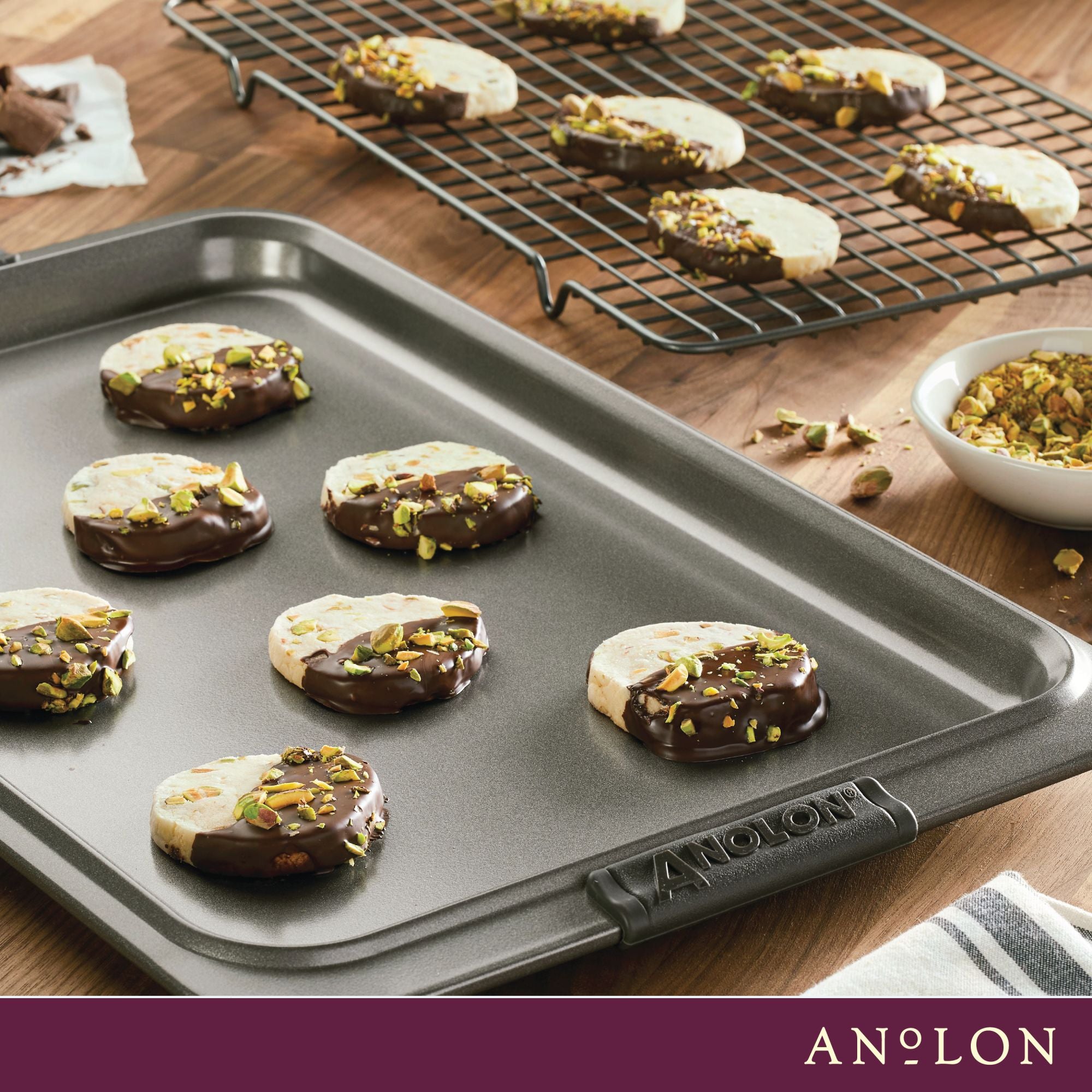 Chocolate-dipped cookies with chopped pistachios rest on the Advanced 11 x 17-Inch Baking Sheet and Cooling Rack Set, with a bowl of pistachios nearby. The Anolon logo appears at the bottom of this elegant scene.