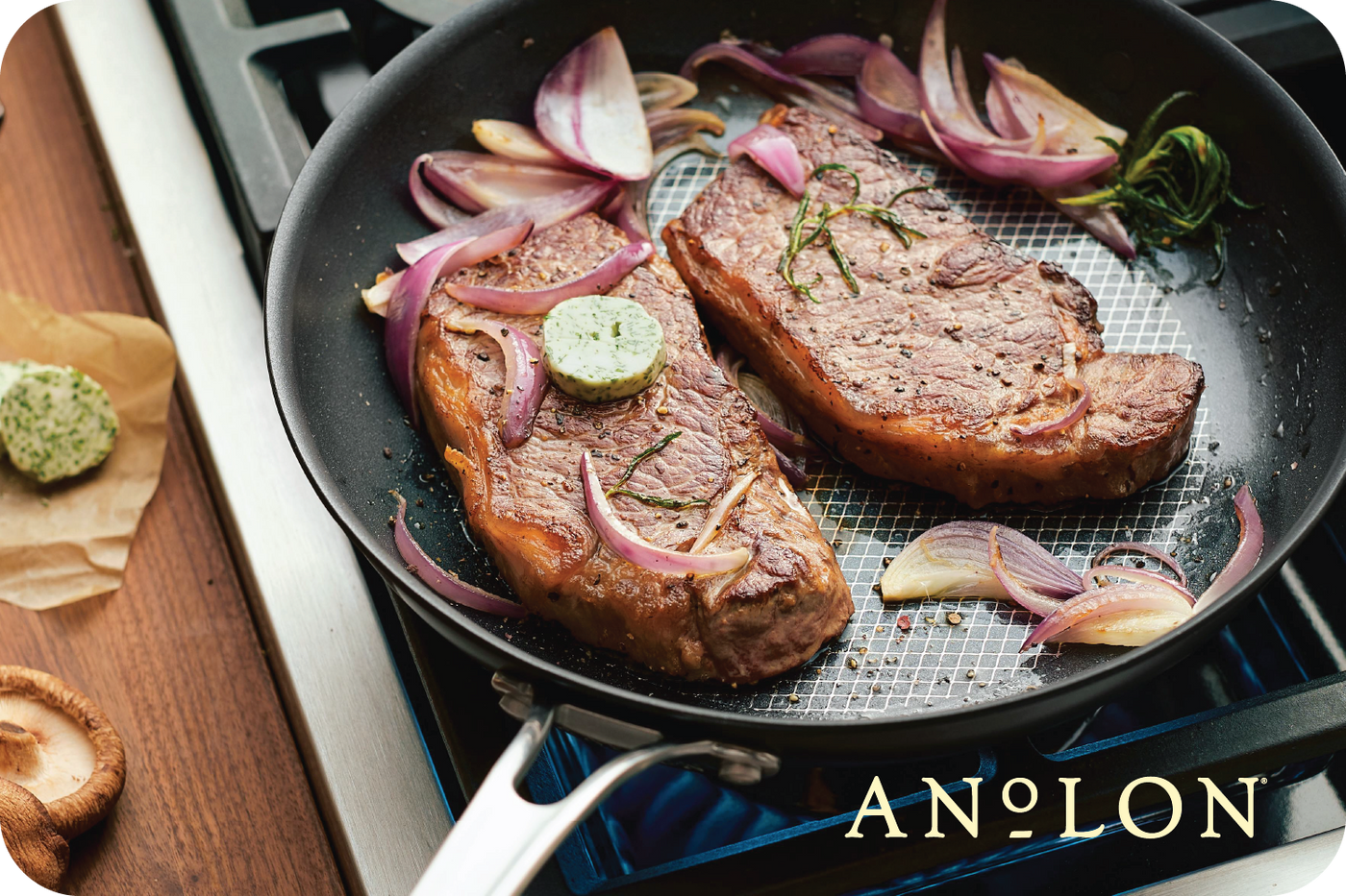 Two seasoned steaks sizzle in an Anolon pan, surrounded by slices of red onion and garlic. A pat of herb butter melts luxuriously on top, filling the kitchen with a mouthwatering aroma. Picture yourself using an Anolon.com Gift Card to recreate this delicious scene at home.
