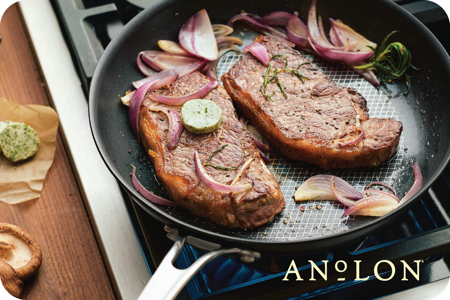 Two seasoned steaks sizzle in an Anolon pan, surrounded by slices of red onion and garlic. A pat of herb butter melts luxuriously on top, filling the kitchen with a mouthwatering aroma. Picture yourself using an Anolon.com Gift Card to recreate this delicious scene at home.