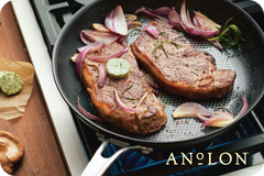 Two seasoned steaks sizzle in an Anolon pan, surrounded by slices of red onion and garlic. A pat of herb butter melts luxuriously on top, filling the kitchen with a mouthwatering aroma. Picture yourself using an Anolon.com Gift Card to recreate this delicious scene at home.