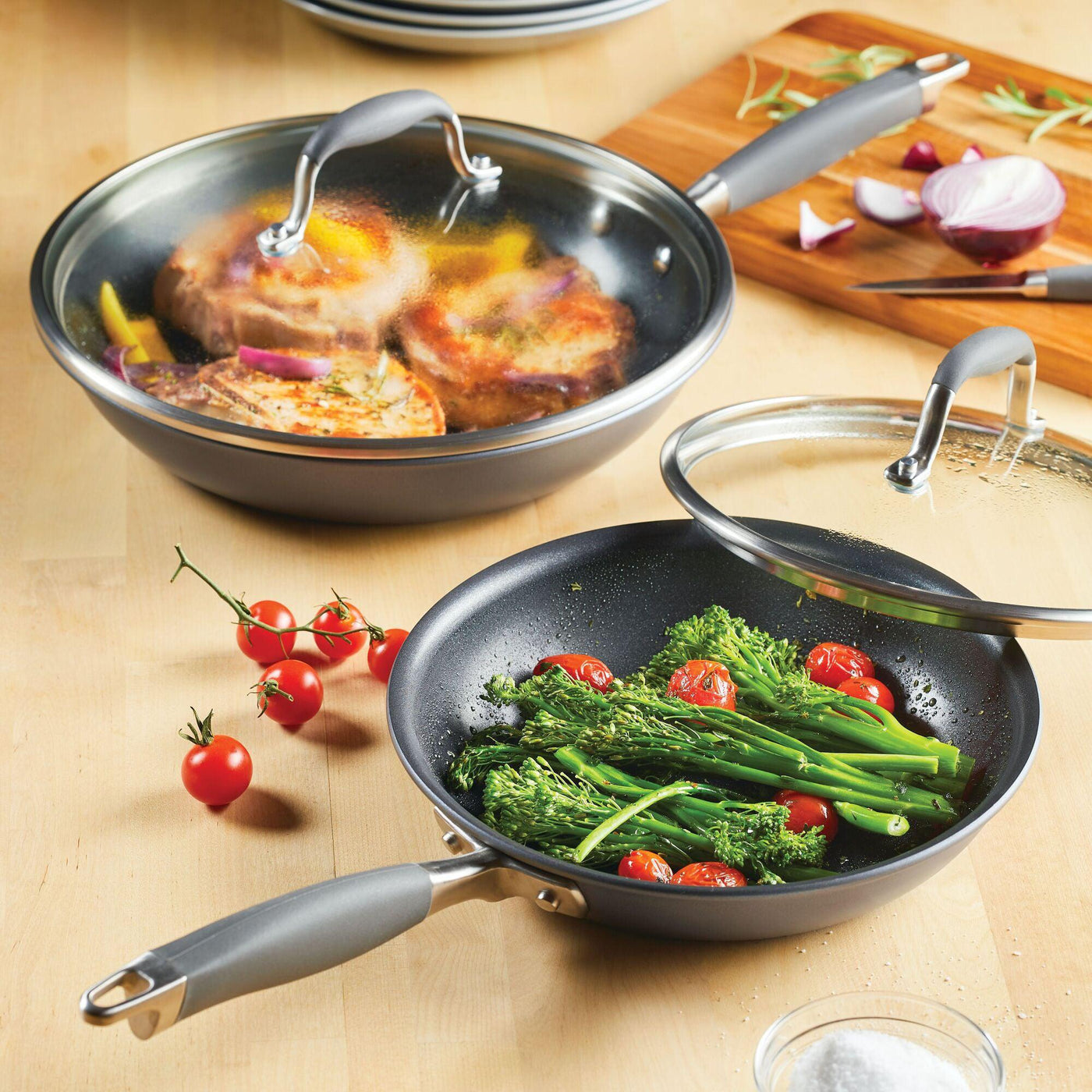 On a wooden table, two skillets from the Advanced Home 4-piece Skillet and Lid Set are featured: one skillet contains cooked meat and onions covered by a tempered glass lid, while the other holds cherry tomatoes and broccolini, partially covered. Nearby is a cutting board with sliced onion, garlic cloves, and some vibrant red cherry tomatoes.