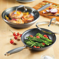 On a wooden table, two skillets from the Advanced Home 4-piece Skillet and Lid Set are featured: one skillet contains cooked meat and onions covered by a tempered glass lid, while the other holds cherry tomatoes and broccolini, partially covered. Nearby is a cutting board with sliced onion, garlic cloves, and some vibrant red cherry tomatoes.
