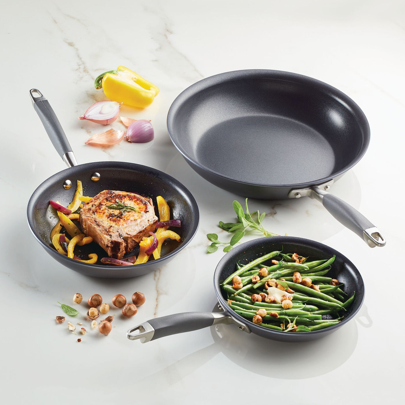 Three frying pans from the Advanced Home 3-Piece Nonstick Frying Pan Set, known for their hard-anodized durability, rest on a marble surface: one with a salmon steak and bell peppers, another with green beans and hazelnuts, and an empty wok. Ingredients such as shallot, pepper, and fresh herbs surround the pans.