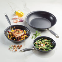 Three frying pans from the Advanced Home 3-Piece Nonstick Frying Pan Set, known for their hard-anodized durability, rest on a marble surface: one with a salmon steak and bell peppers, another with green beans and hazelnuts, and an empty wok. Ingredients such as shallot, pepper, and fresh herbs surround the pans.
