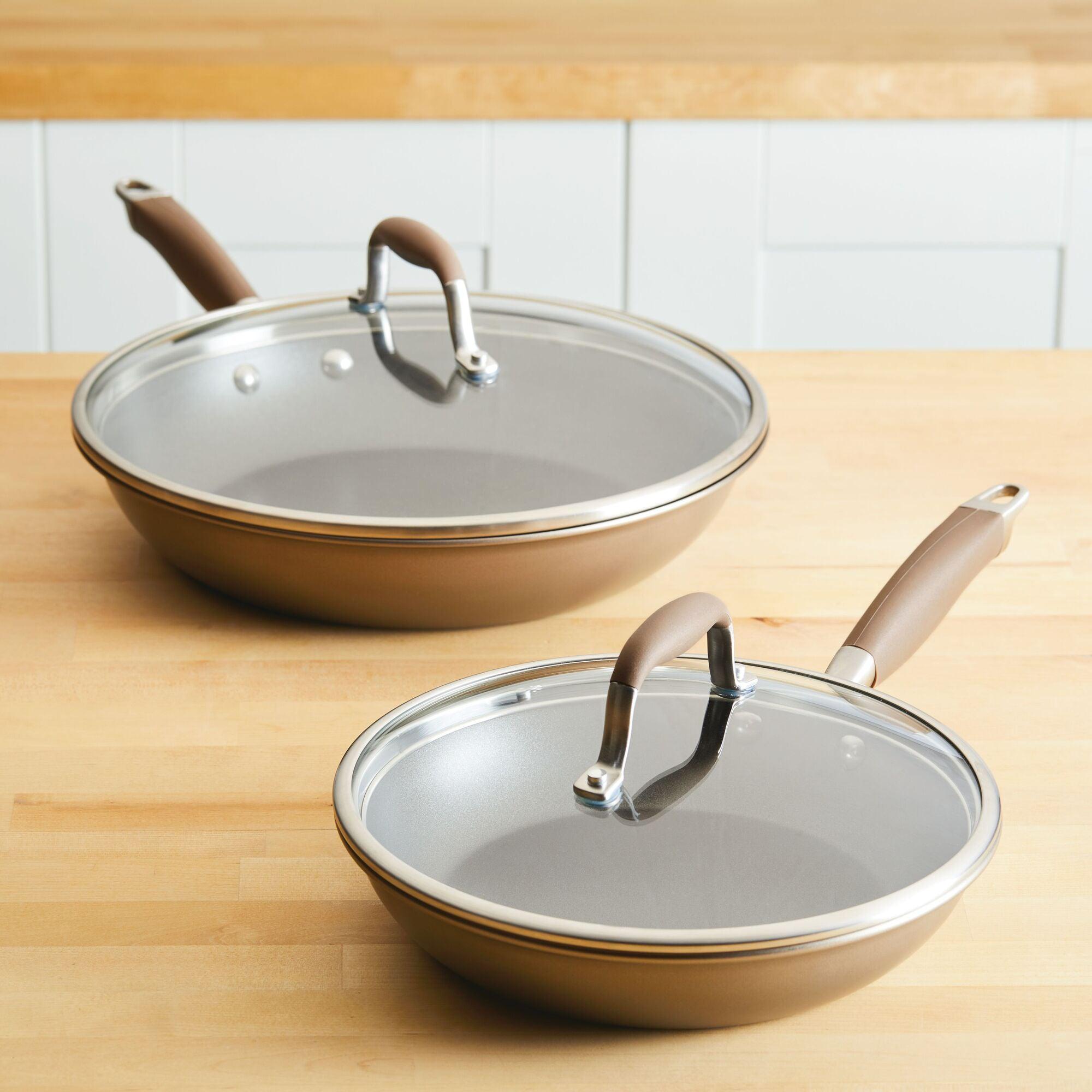 A 4-piece Skillet and Lid Set by Advanced Home, featuring two non-stick frying pans with Infinity Slide surfaces and tempered glass lids, rests on a wooden kitchen countertop. The pans boast brown handles and a metallic exterior, complementing the light-colored kitchen backdrop.