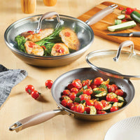 On a wooden surface, two frying pans from the Advanced Home 4-piece Skillet and Lid Set showcase a delightful culinary scene: one holds sautéed cherry tomatoes and zucchini, while the other is filled with cooked chicken, greens, and cherry tomatoes. Their hard-anodized durability and Infinity Slide nonstick coating ensure spotless cooking experiences, with tempered glass lids conveniently placed for effortless monitoring.