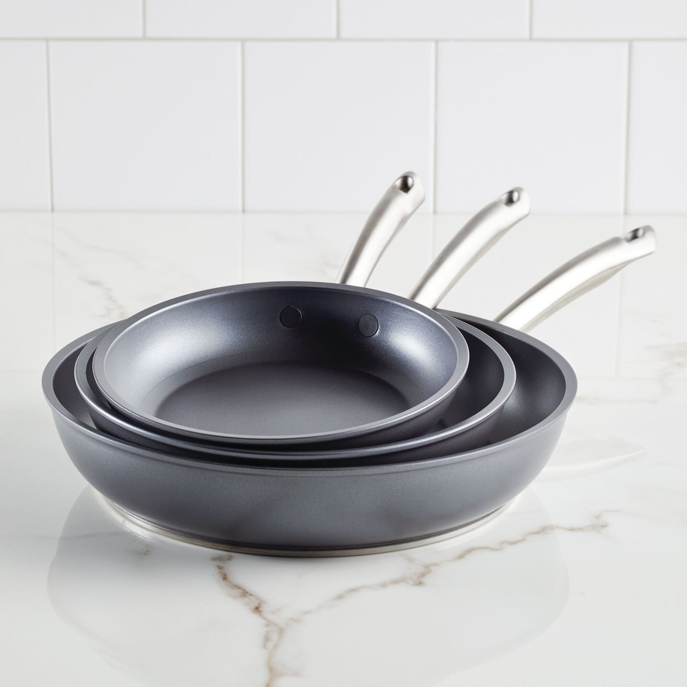 An Accolade 3-Piece Nonstick Frying Pan Set with shiny metal handles, designed using Precision Forge™ construction, is neatly nested on a white marble countertop. White subway tiles provide the backdrop.
