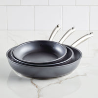 An Accolade 3-Piece Nonstick Frying Pan Set with shiny metal handles, designed using Precision Forge™ construction, is neatly nested on a white marble countertop. White subway tiles provide the backdrop.