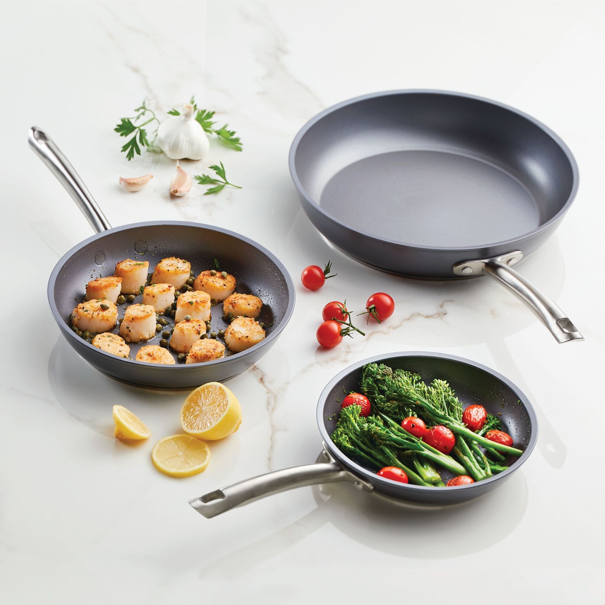On a marble countertop, three Accolade 3-Piece Nonstick Frying Pan Set are beautifully showcased: one holds cooked scallops, another contains roasted broccolini with cherry tomatoes, and the third pan is empty. Surrounding the pans, you can see garlic, parsley, cherry tomatoes, and lemon wedges. These durable pans are oven-safe and can withstand temperatures up to 500°F.
