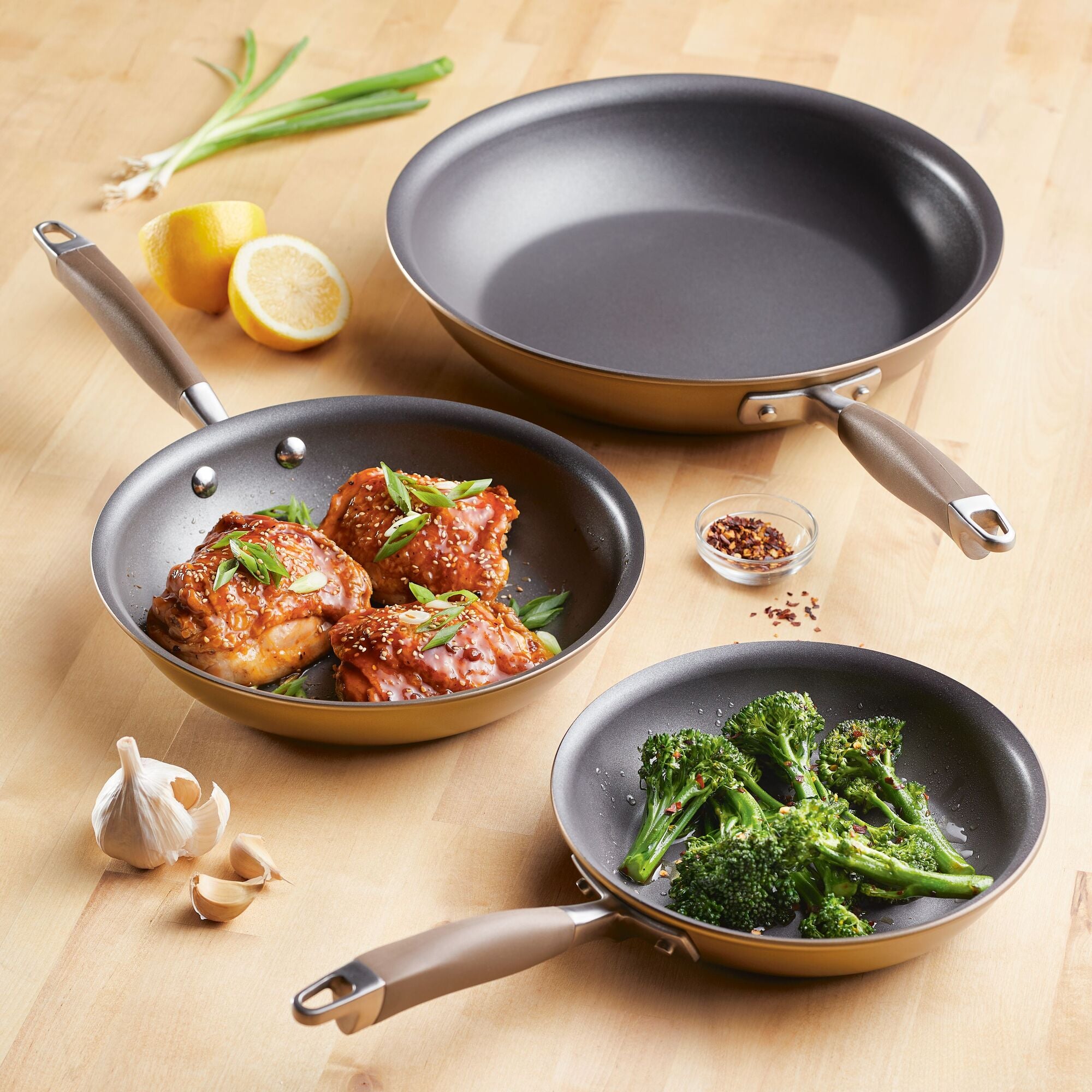 On a wooden surface, three Advanced Home Infinity Slide nonstick frying pans from the 3-Piece Nonstick Frying Pan Set are displayed. One pan is filled with chicken thighs in sauce and herbs, another holds sautéed broccoli, and the third one is empty. Nearby, there are ingredients such as lemon, garlic, and red pepper flakes—ideal for complementing your nonstick cooking experience.