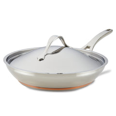 The Nouvelle Stainless 12-Inch Frying Pan with Lid features a sleek, curved handle and a gleaming, reflective surface, complemented by its copper base. It is elegantly displayed against a plain white background.