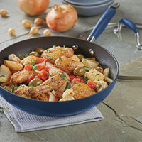 The Advanced 12-Inch Covered Ultimate Pan, shown in blue and filled with chicken, potatoes, and veggies on a gray napkin with onions and garlic nearby, is perfect for easy one-pan meals.