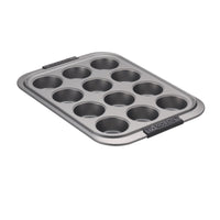 The Advanced Muffin Pan with Lid, designed for baking delicious treats, features a 12-cup nonstick surface and is crafted from heavy gauge steel with a metallic finish. It includes two black silicone grips on the sides for easy handling. The pan is displayed at a slight angle against a white background.