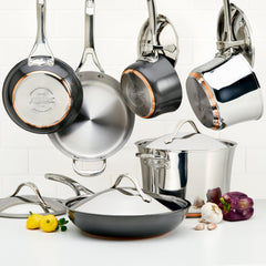 An assortment of stainless steel pots and pans from the Nouvelle Luxe 11-Piece Mixed Metals Cookware Set, renowned for their nonstick performance, hangs gracefully against a white tiled wall accentuated by copper cookware. A handful are placed on a white surface accompanied by lemons, garlic, a purple bell pepper, and a red onion.