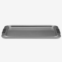 The Cookie Sheet with Silicone Grips by Advanced is a rectangular non-stick pan featuring dark gray silicone grips on each side, contrasting with a light gray background. It offers even heat distribution and has a flat surface with raised edges designed for optimal baking and easy handling.