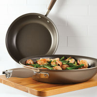 The Advanced Home 10.25 & 12.75-Inch Frying Pan Set, made of hard-anodized aluminum, is shown resting on a wooden cutting board; one pan contains cooked shrimp and green beans, set against a white tiled kitchen wall.