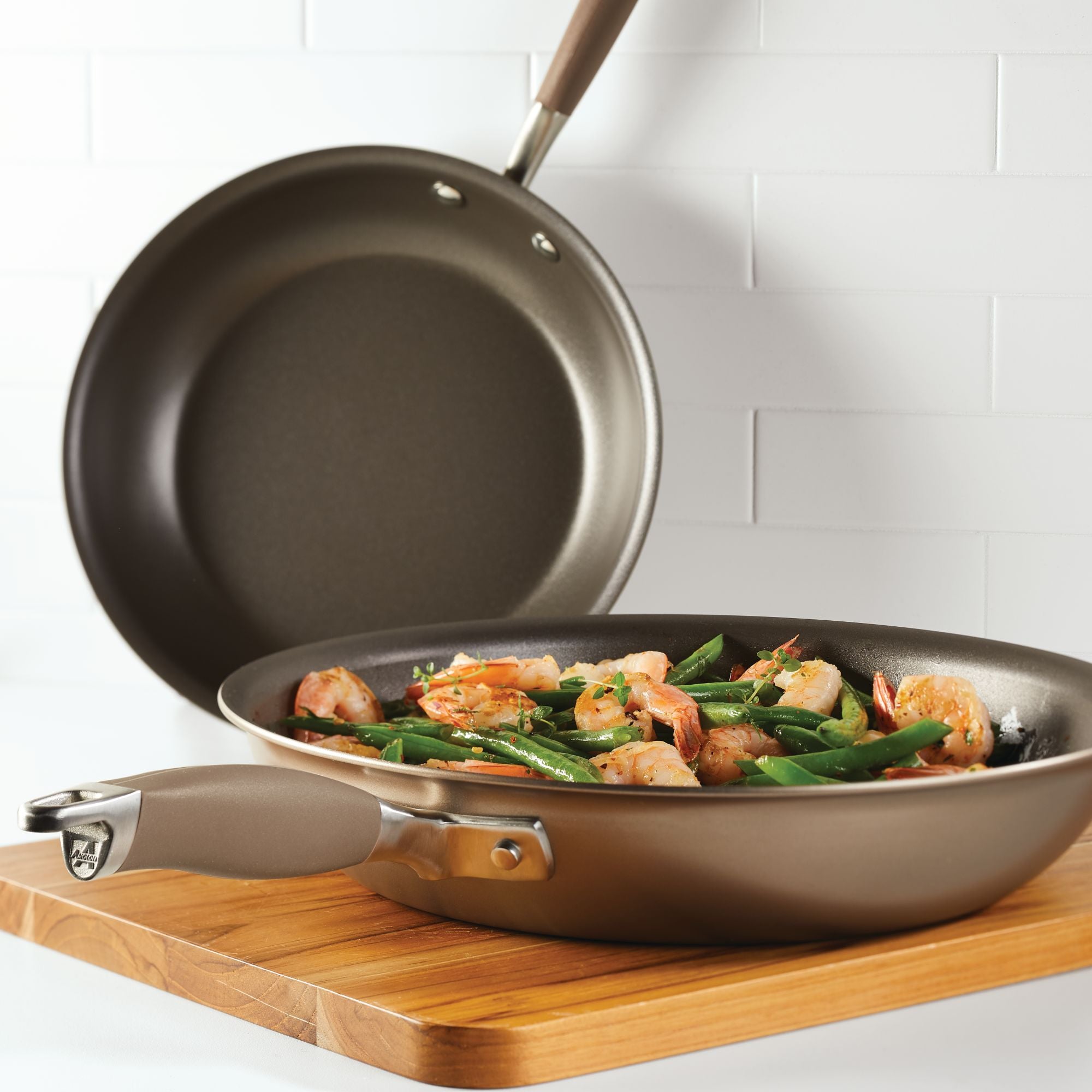 The Advanced Home 10.25 & 12.75-Inch Frying Pan Set, made of hard-anodized aluminum, is shown resting on a wooden cutting board; one pan contains cooked shrimp and green beans, set against a white tiled kitchen wall.